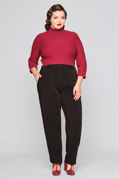 Curvy brunette woman standing with one hand in her pocket wearing burgundy high heels, black trousers and a burgundy lattice knitted jumper