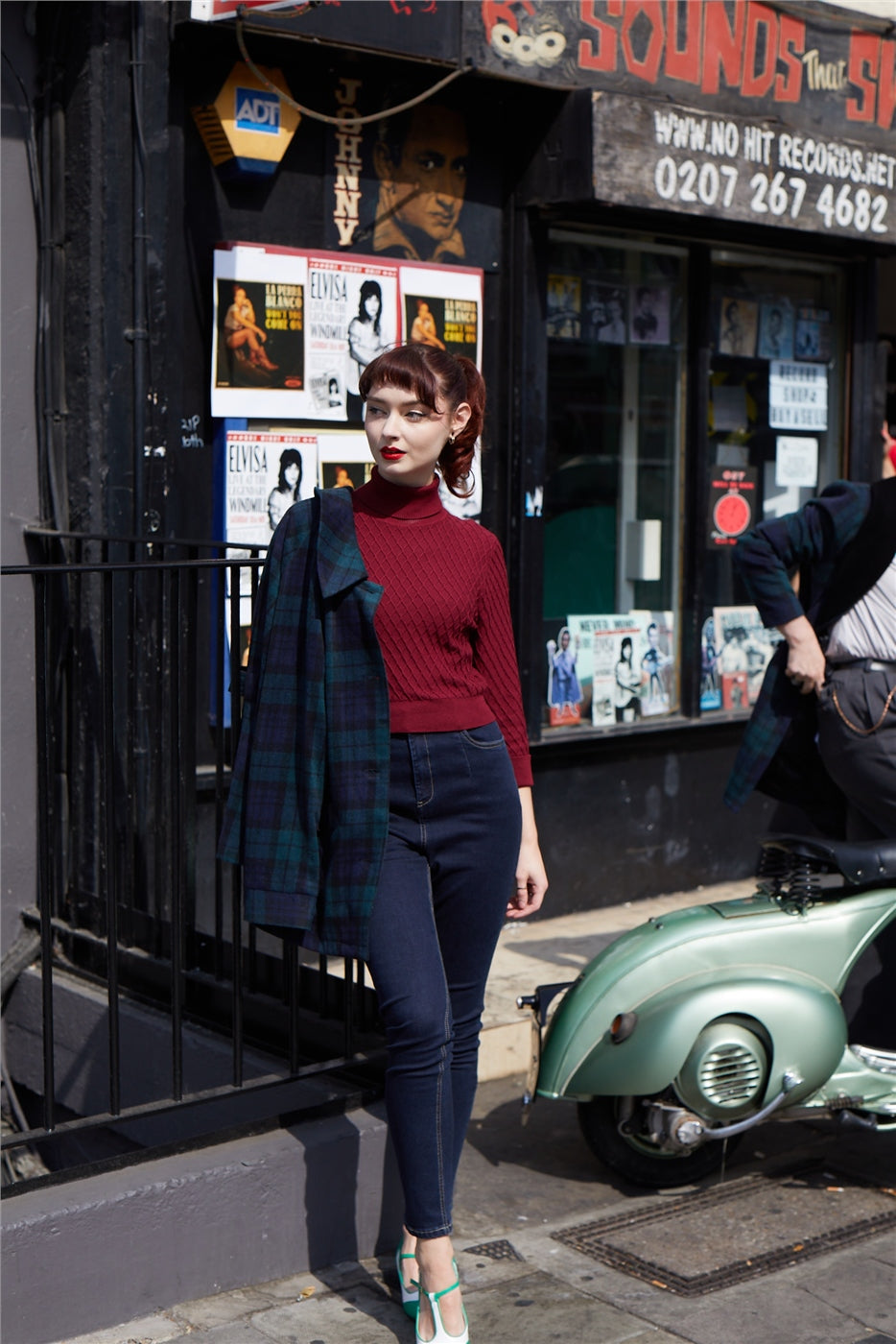 Young brunette woman with her coat over one shoulder standing outside a shop in London wearing high waisted skinny trousers and burgundy jumper