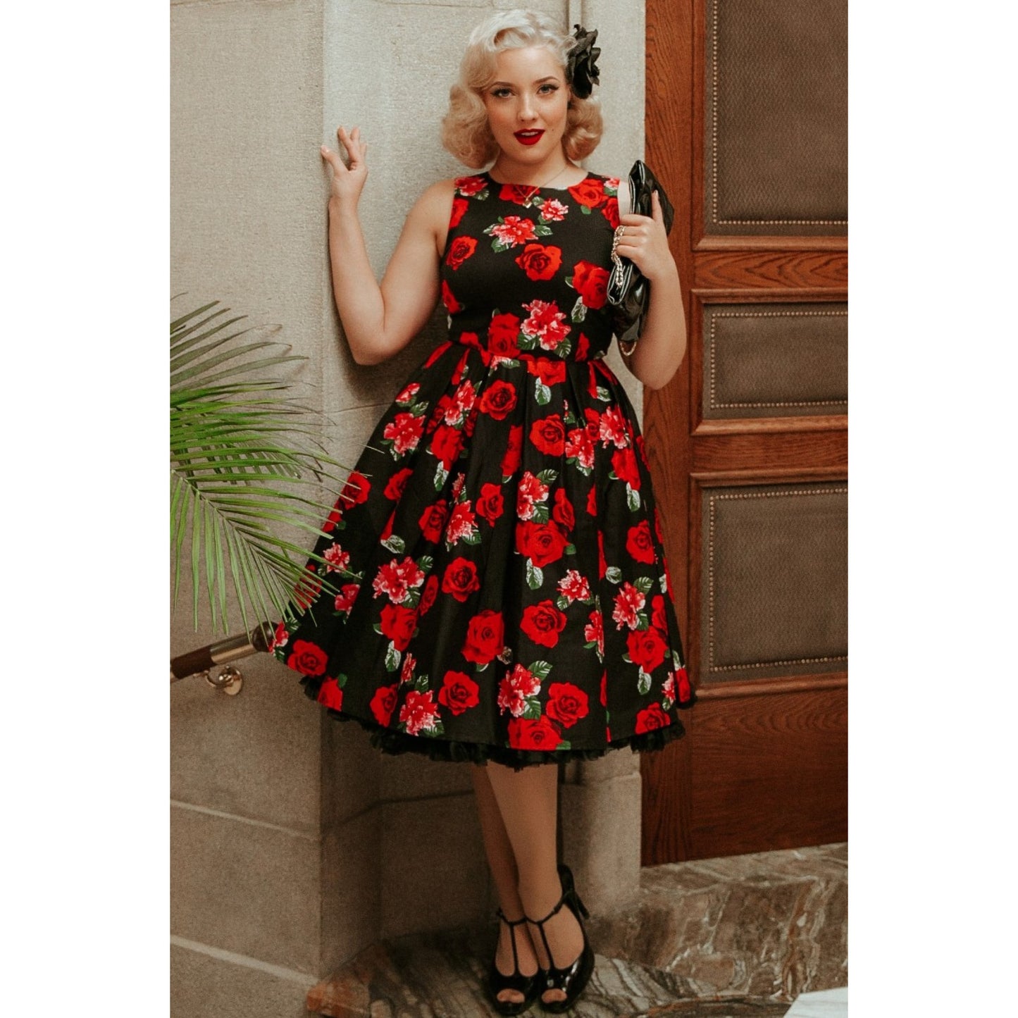 Blonde woman wearing red lipstick standing in a stone hallway wearing the Annie red roses dress with a black petticoat underneath and black T Bar high heels