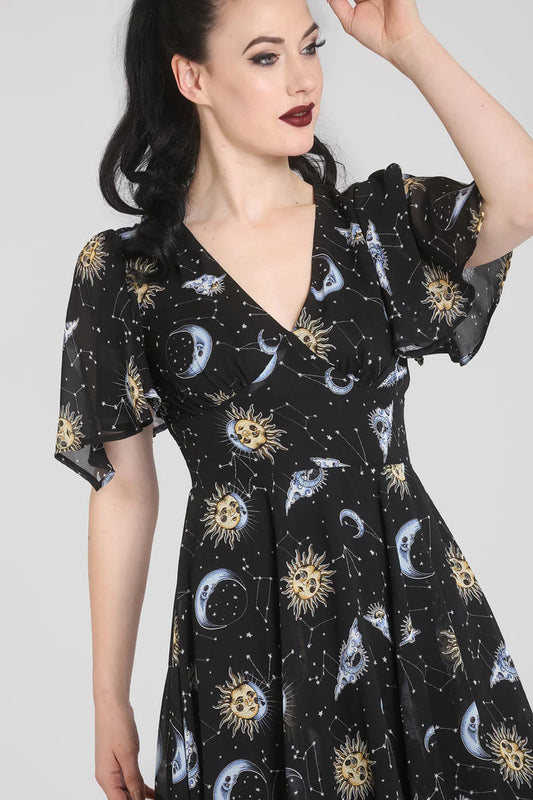 Dark haired girl with dark red lipstick wearing a black moon, stars and constellations print dress with short fluttery sleeves and V neck standing with one hand raised to her face.