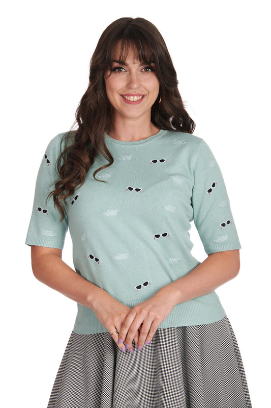 Happy, smiling woman with long wavy brown hair wearing a light blue 3/4 length sleeve knit fabric top with embroidered sunglasses and wave motif all over