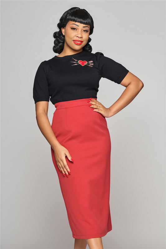 Tall elegant woman with black curled hair standing with one hand on her hip wearing a red high waisted pencil skirt and a black T-shirt top with a heart and arrows motif on the right side of the chest.