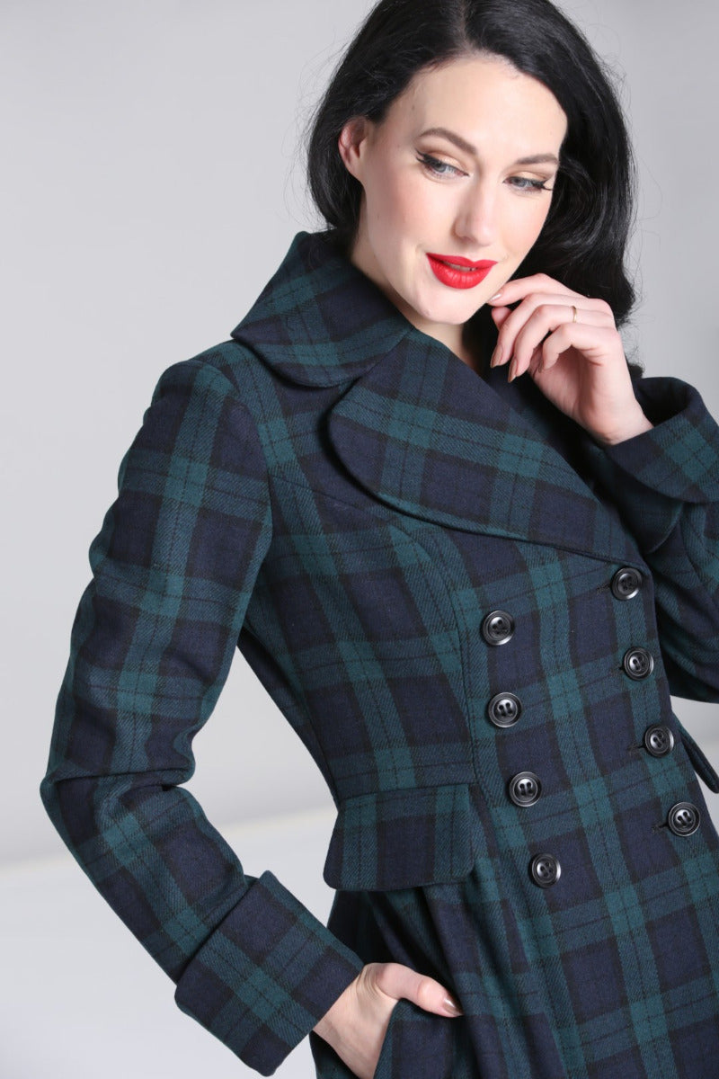 Dark haired vintage style woman with one hand resting on her cheek and the other tucked into her coat pocket. The Coat is navy and green tartan with a wide collar and black buttons at the front