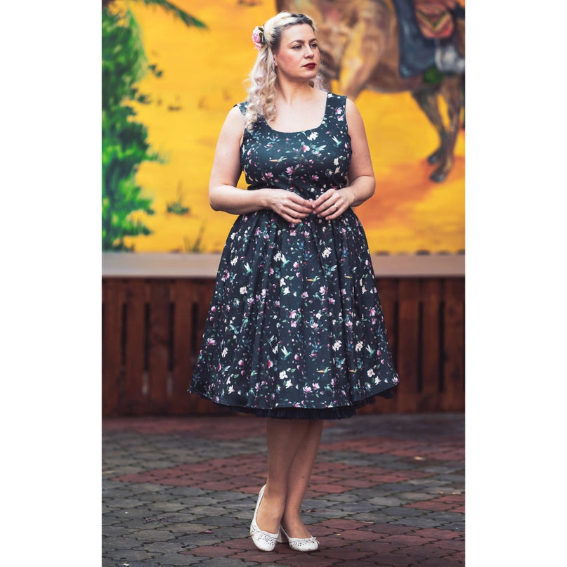 Model wearing the Amanda Swing Dress in Navy Blue Hummingbird Print by Dolly & Dotty. The dress features a floral and hummingbird print on a navy background, with a sleeveless design, fitted bodice, and a full swing skirt. The model stands against a colorful mural background, showcasing the vintage-inspired elegance of the dress.
