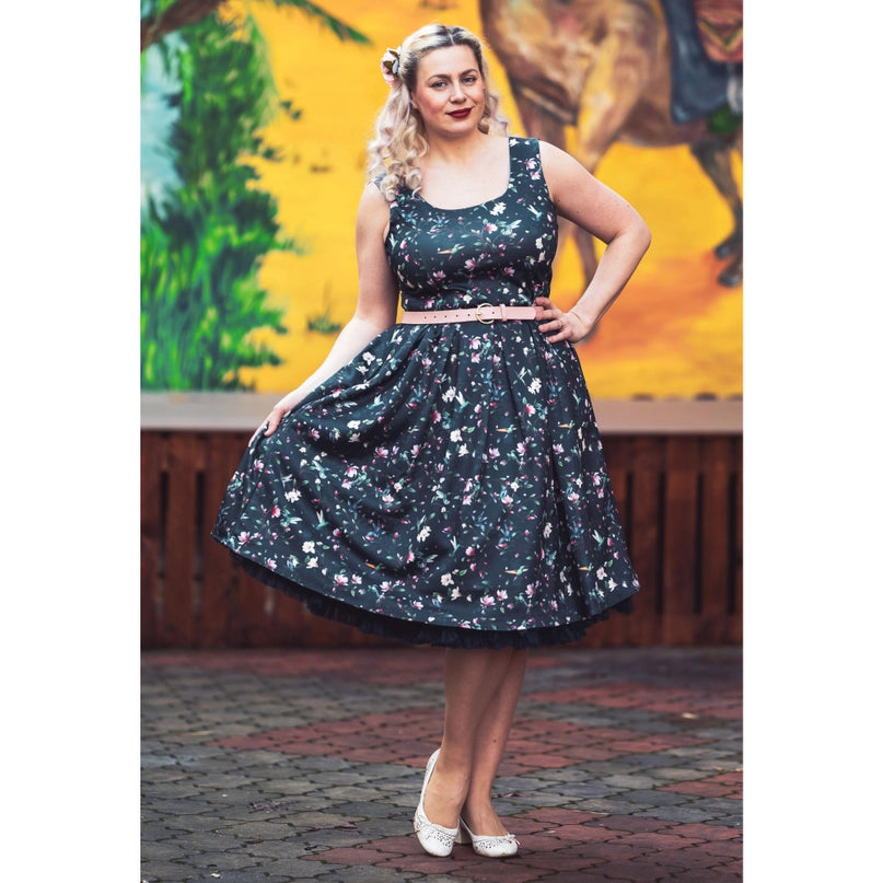 Model wearing the Amanda Swing Dress in Navy Blue Hummingbird Print by Dolly & Dotty, styled with a light pink belt to accentuate the waist. The dress features a vibrant floral and hummingbird print on a navy background, with a sleeveless design, square neckline, and a full, vintage-inspired swing skirt. The model stands outdoors against a colorful mural, giving the look a retro and elegant vibe.