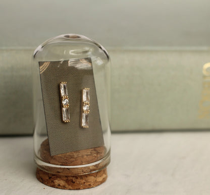 Gold and crystal earrings inside a glass dome on a light gray surface.
