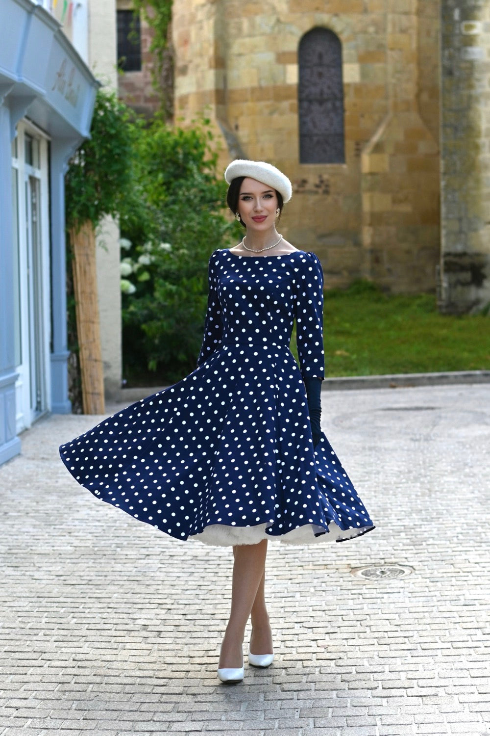 Woman in a blue polka dot dress wearing a white beret and pearl necklace standing on a street with a stone building in the background 