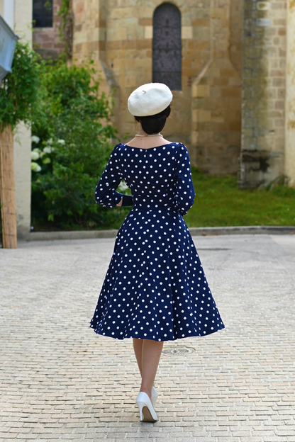 Woman wearing a navy blue dress with white polka dots and a white hat, standing in front of a stone building.