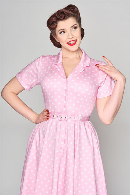 Brunette woman with vintage style victory rolls in her hair standing with one hand on her hip and the other resting on her shoulder. She wear the Caterina pink polka dot dress by Collectif.