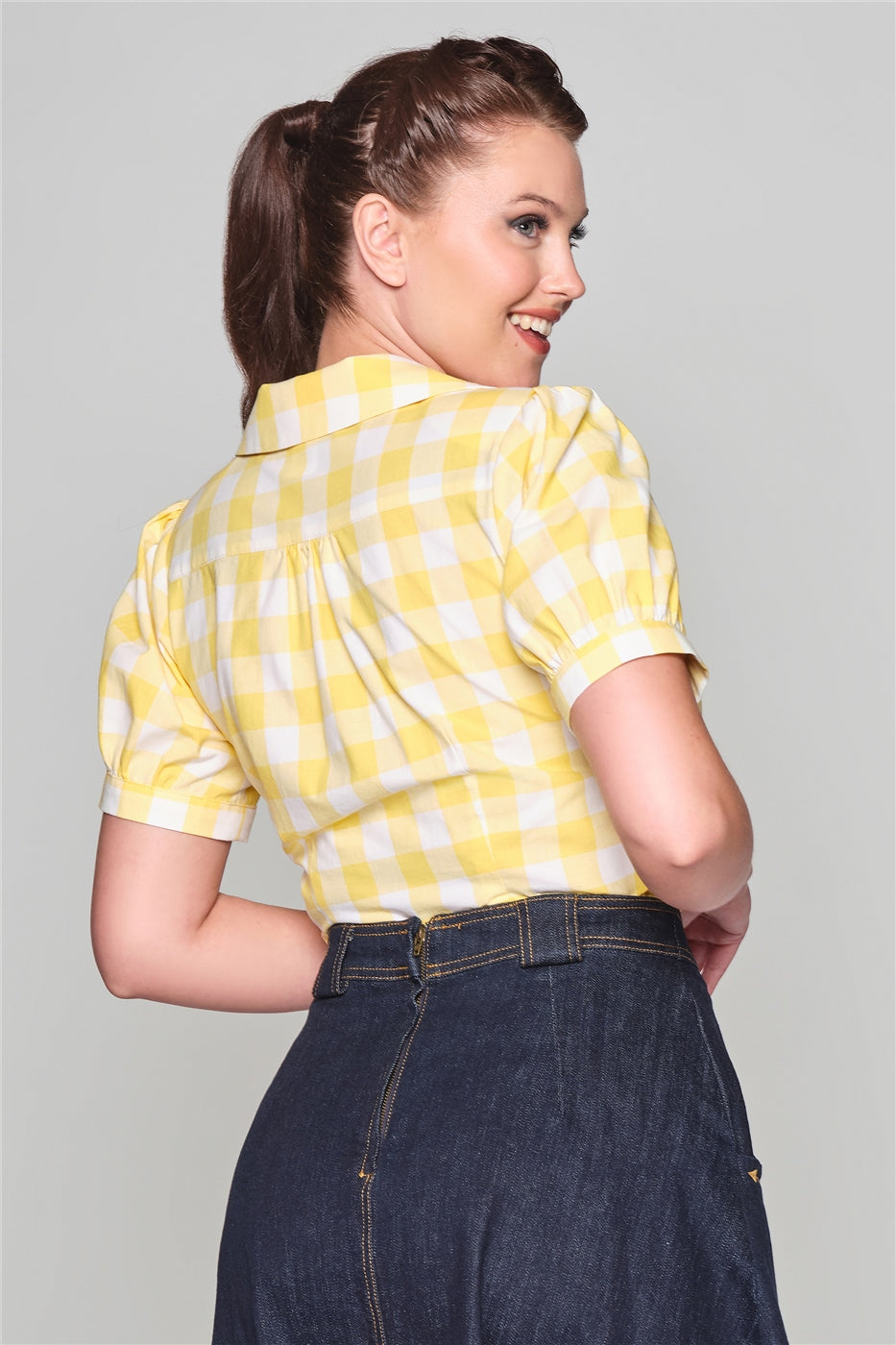 Happy, smiling girl looking over her shoulder with her hair tied back in a ponytail. She is wearing a denim skirt and yellow gingham short sleeved blouse. 