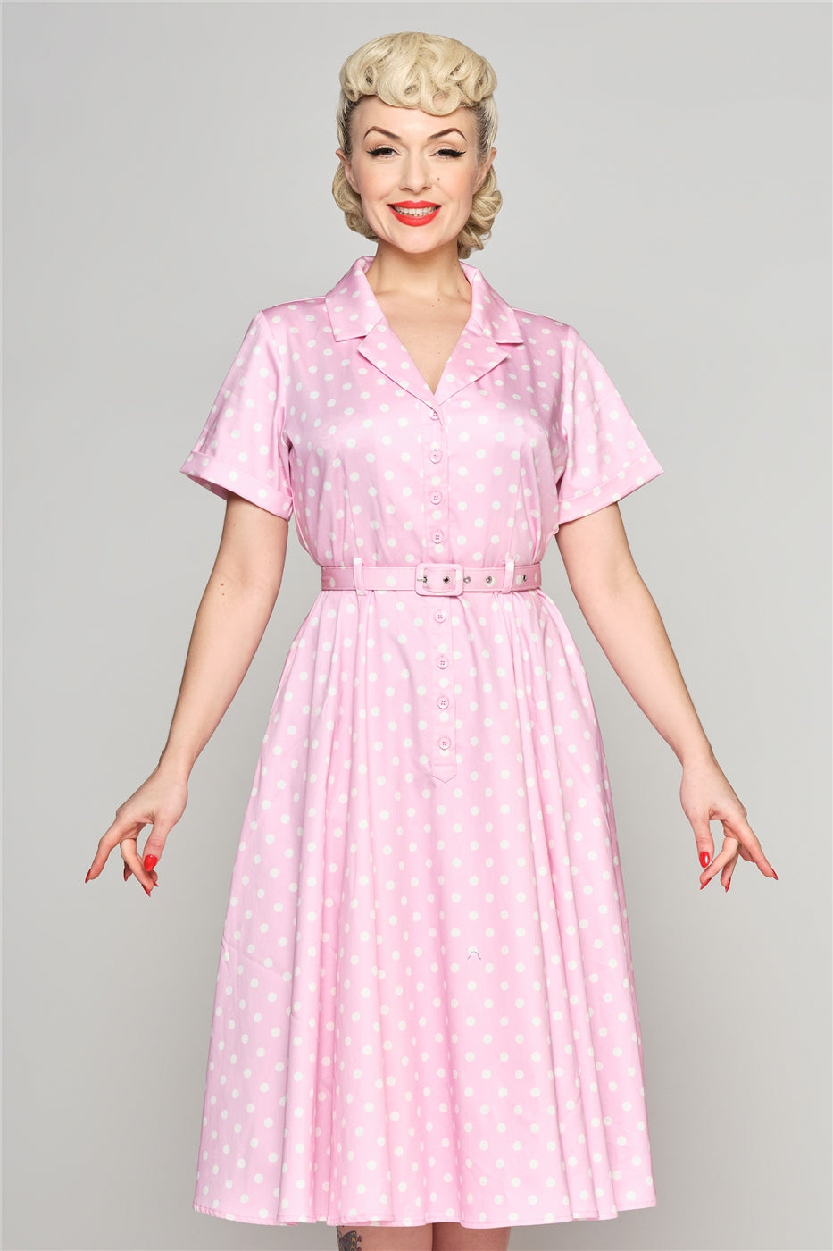 Elegant blonde woman with her hair in an up-do standing wearing a pink polka dot shirt dress with a matching belt.