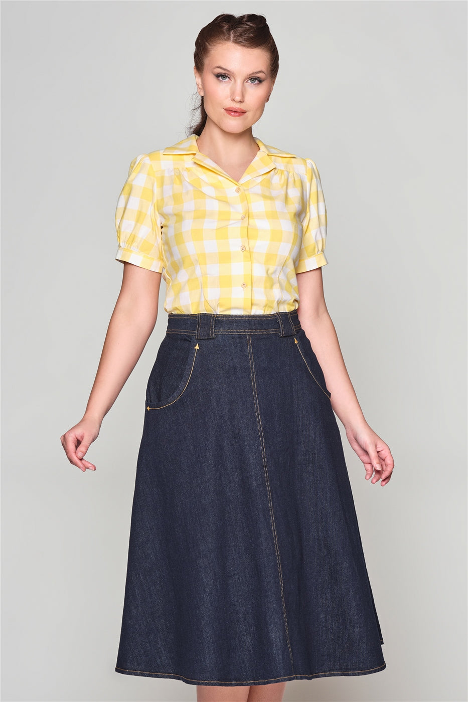 Young woman with a ponytail standing with her arms by her sides wearing the Luana yellow gingham blouse and a denim mid length A-line skirt.