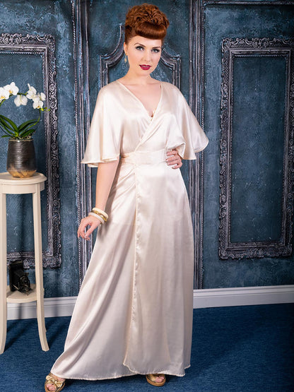 Model wearing an elegant 1930s-inspired peach satin lounging robe by What Katie Did, styled with vintage gold accessories, standing against a classic blue and silver wall backdrop with an orchid plant on a side table.