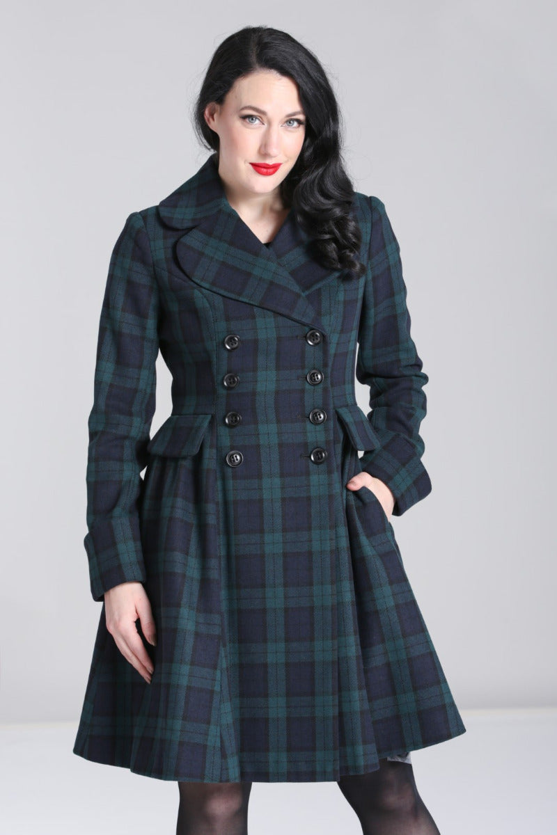 Smiling woman standing with one hand in her coat pocket wearing red lipstick, shee black tights and a navy and green tartan swing coat