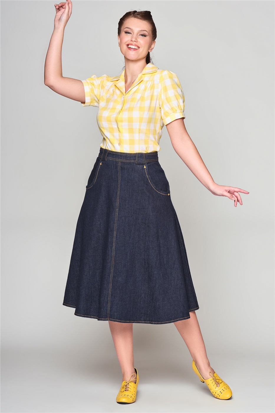 Happy woman with a radiant smile with one arm up in the air and the other by her side wearing a denim skirt, yellow gingham 40s style blouse and yellow heeled shoes.