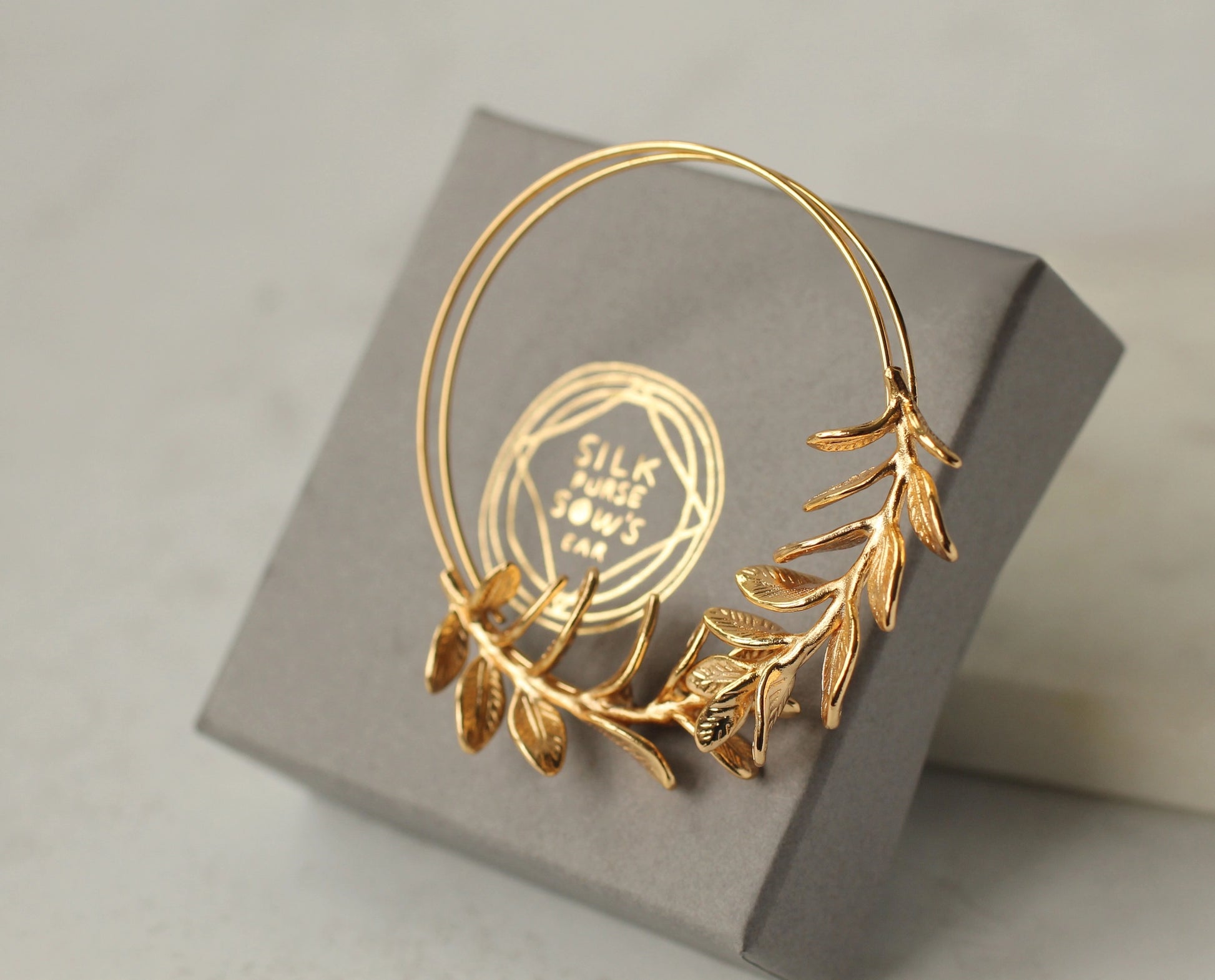 Gold hoop earring with leaf details on a gray box with 'Silk Purse Sow's Ear' branding.