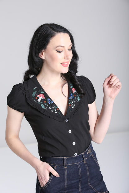 Fair skinned dark haired girl smiling and looking down wearing high waisted blue jeans and a black semi fitted blouse with a woodland embroidery collar