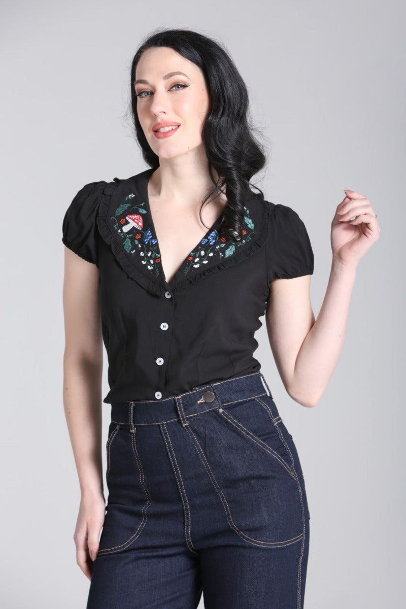 Dark haired model with one hand raised and the other down by her side wearing a semi fitted black blouse with embroidery detail and high waisted denim jeans