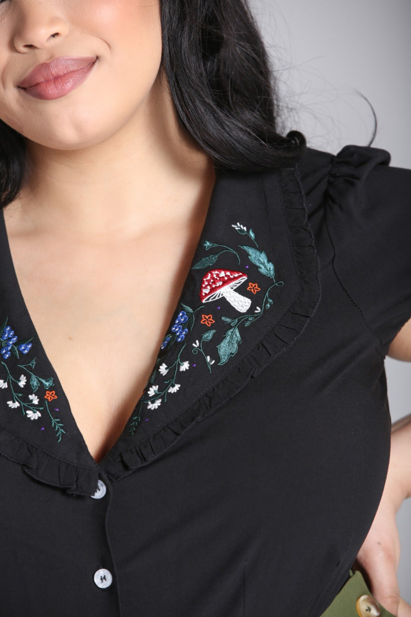 Close up of the collar of a black blouse with embroidered mushrooms, flowers and leaves on it and a frilled edge.