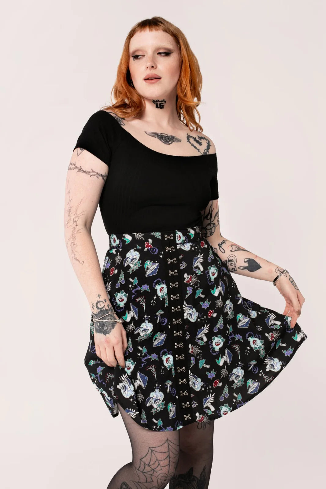 Woman with shoulder length ginger hair and tattoos wearing a black off the shoulder top and a mushroom print mini skirt that falls to just above the knee.