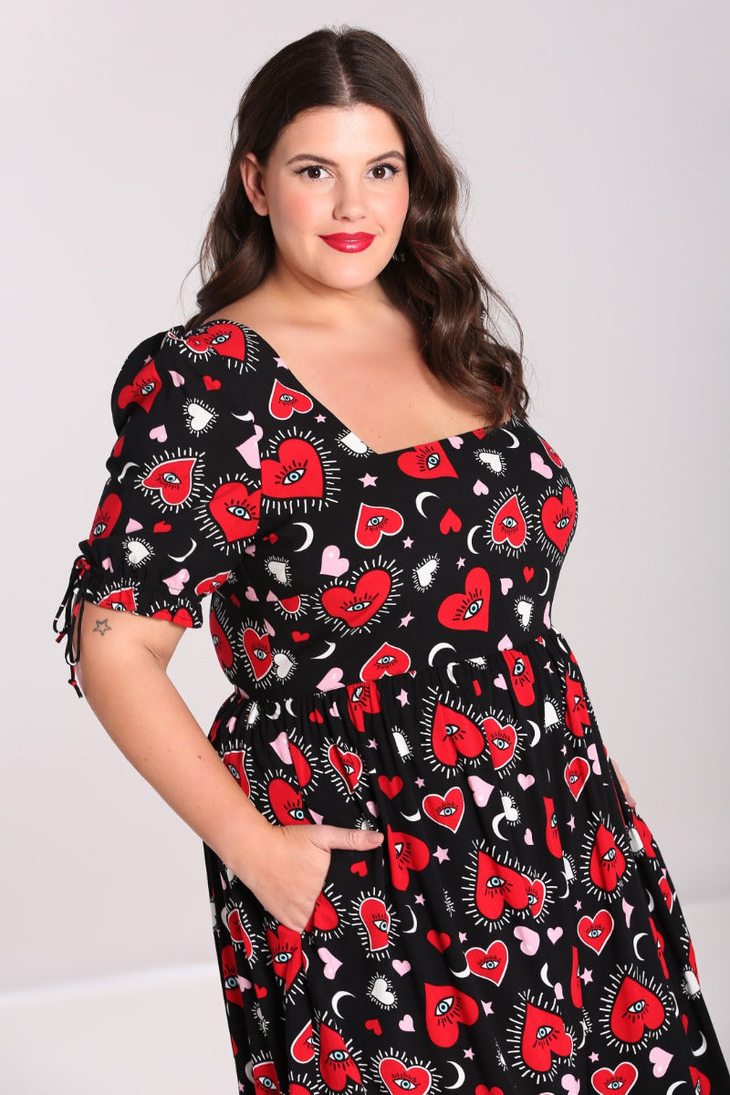 Pretty brunette woman standing with a subtle smile wearing a Heart print black maxi dress with a square neckline and short sleeves. She has her hands in the pockets of the dress.