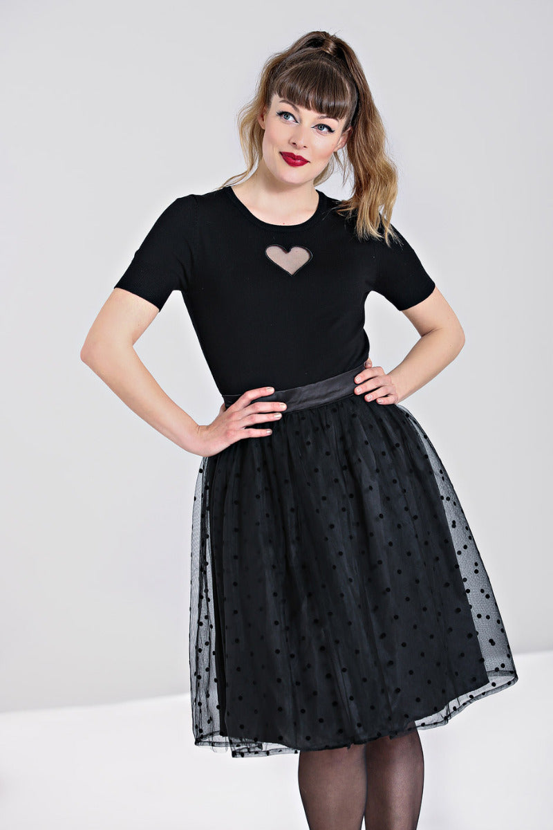 Girl with a ponytail wearing red lipstick standing with her hands on her hips. She is wearing a black t-shirt with a cutout heart and a knee length 50s skirt with black polka dot mesh overlay.