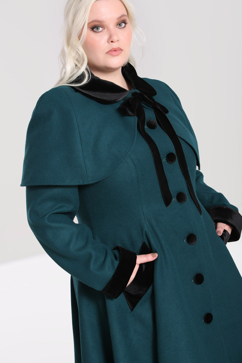Blonde haired plus size model with smoky eye makeup wearing a green cape and 0s style coat  standing with her hands in her coat pockets.  