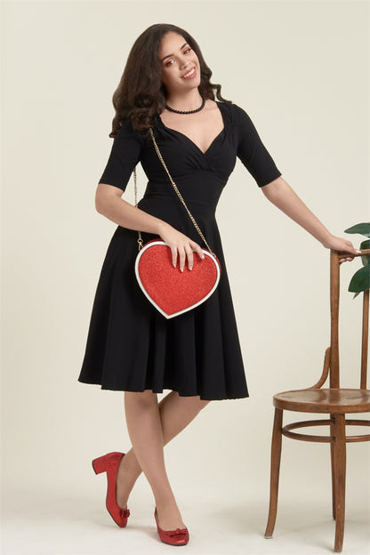 Brown haired girl wearing a black swing dress, black bead necklace, red glittery heeled shoes and a heart shaped glittery red handbag leaning against a wooden chair