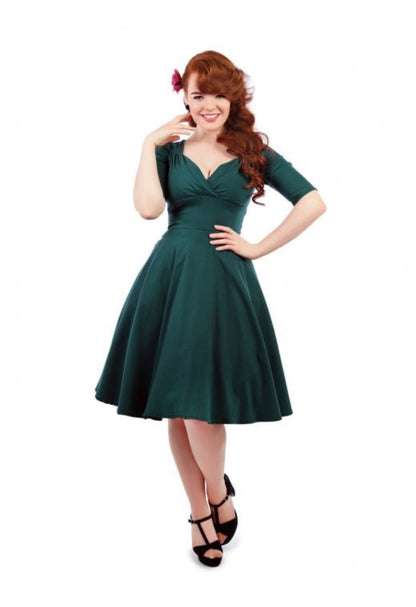50s style fashion model with long curly auburn hair smiling with one hand on her hip and the other raised up to her face wearing a teal swing dress and black high heels