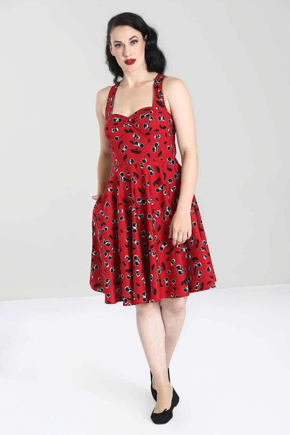 Pinup style dark haired woman with red lipstick wearing a red strappy knee length fit and flare dress with an all over black cherry print.