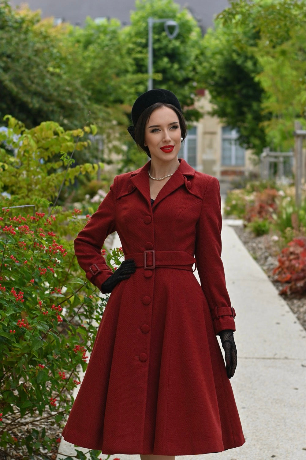 Woman in a red coat standing outdoors with greenery and a building in the background
