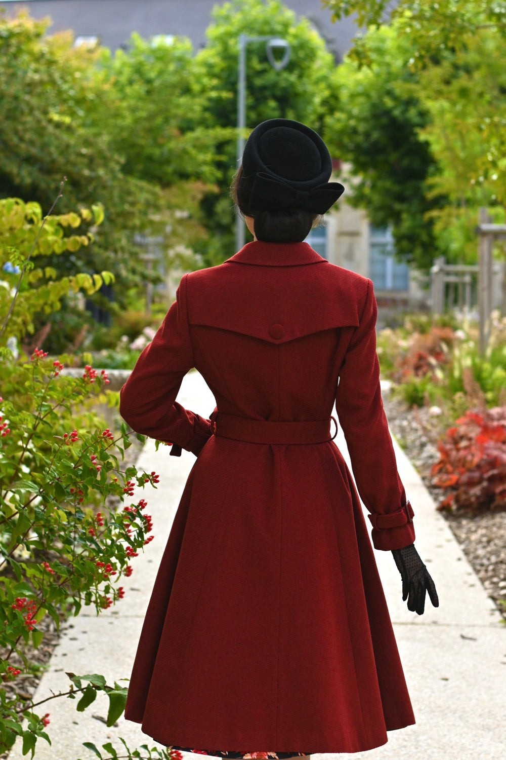 Person wearing a red coat and black hat walking through a garden.