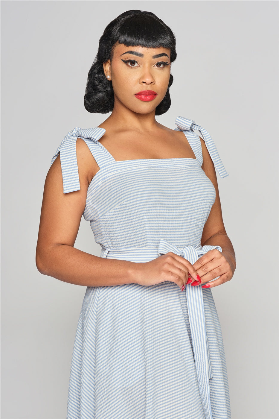 Woman with 50s style makeup wearing a blue and white stripe seersucker dress standing with her hands in front of her waist.