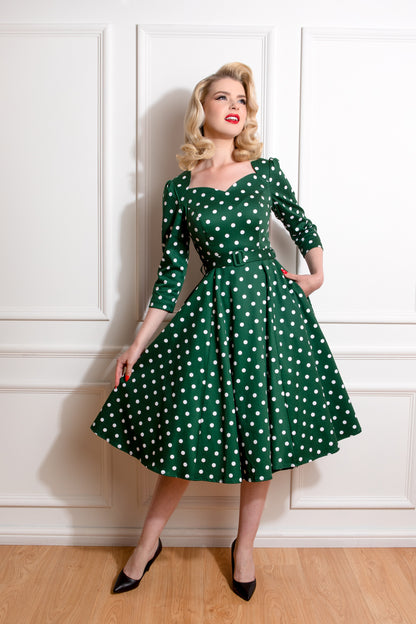 Woman with blonde hair and red lipstick wearing a green dress with white polka dots against a white wall.