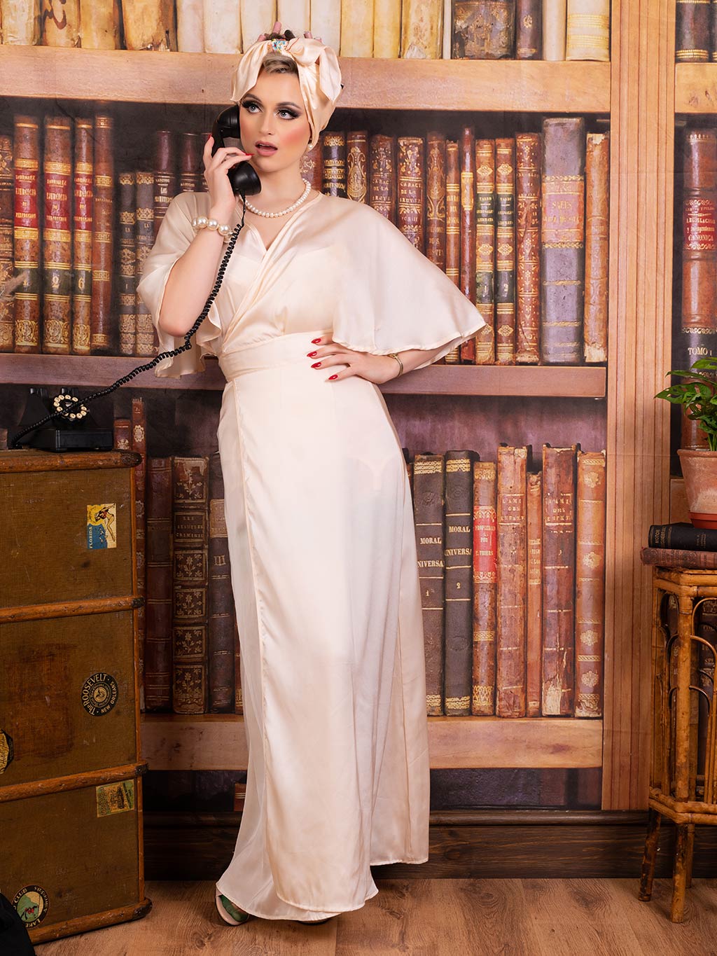 A tall, elegant woman standing in a vintage library with one hand on her hip and the other hand holding a 1950s style telephone to her ear. She wears a floor length satin robe and a hair wrap. and red nail polish