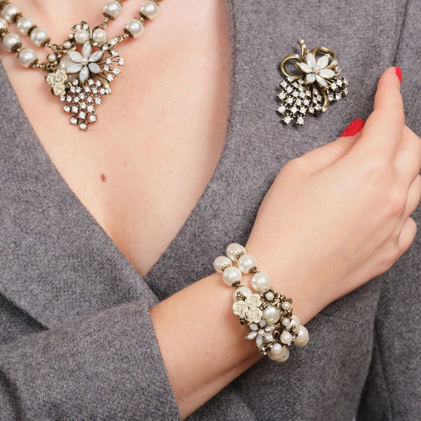 Pearl bracelet, necklace, and brooch on a person wearing a gray outfit.