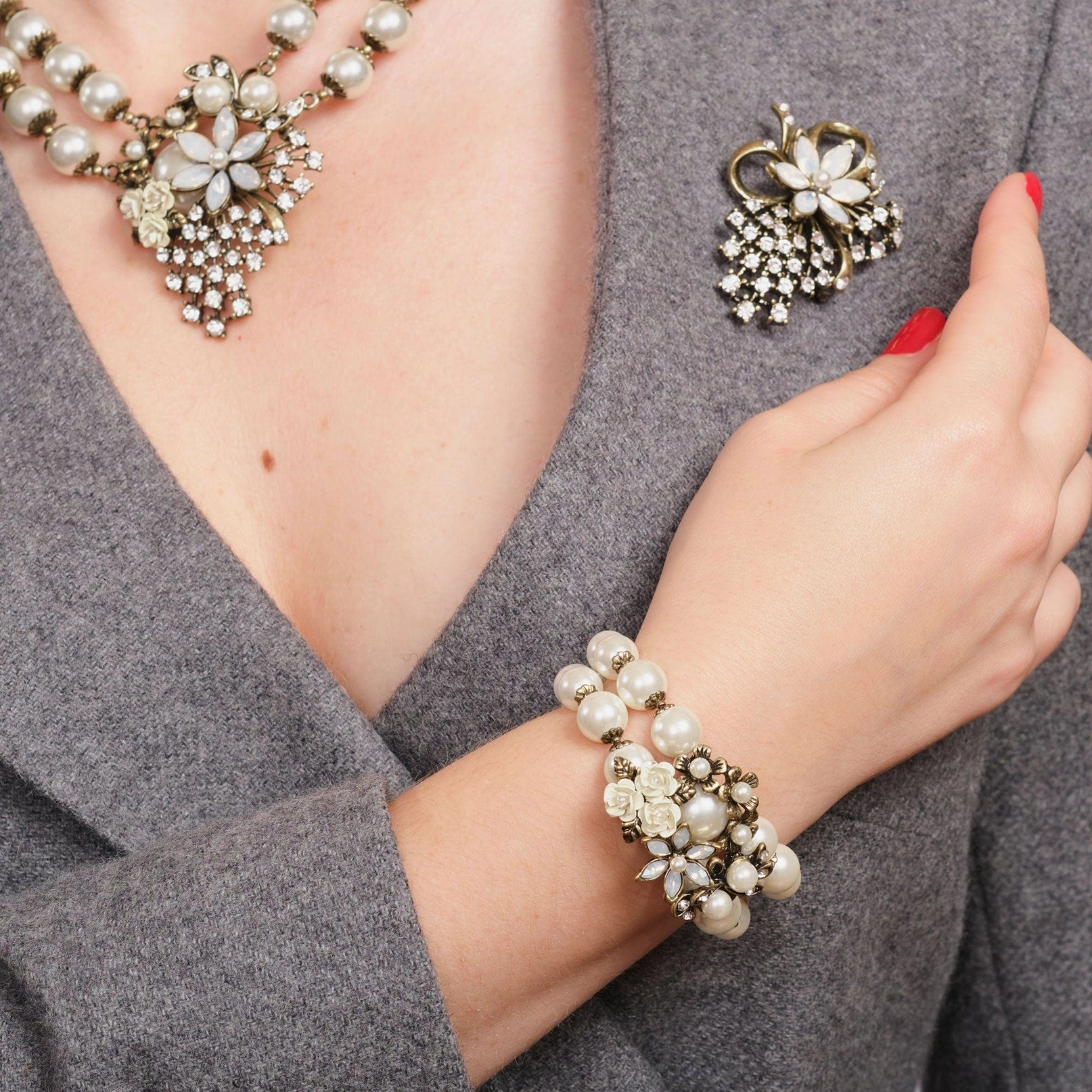 Pearl bracelet, necklace, and brooch on a person wearing a gray outfit.