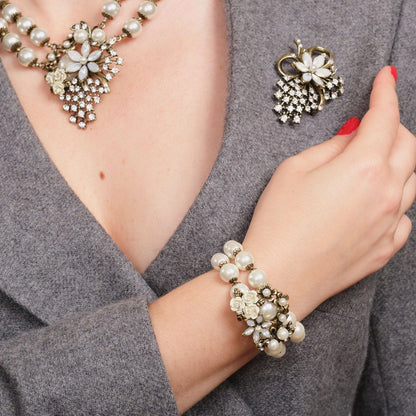 Pearl bracelet, necklace, and brooch on a person wearing a gray outfit.