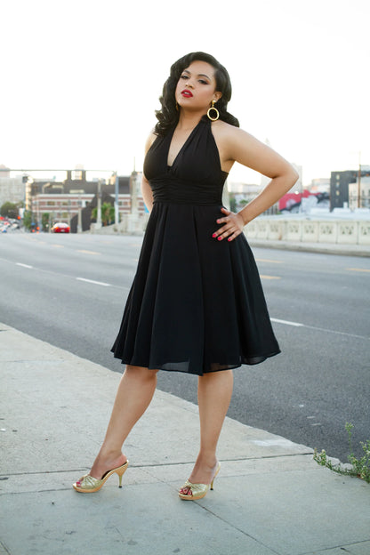 Woman in a black halter neck knee length dress standing on a city street with one hand on her hip.