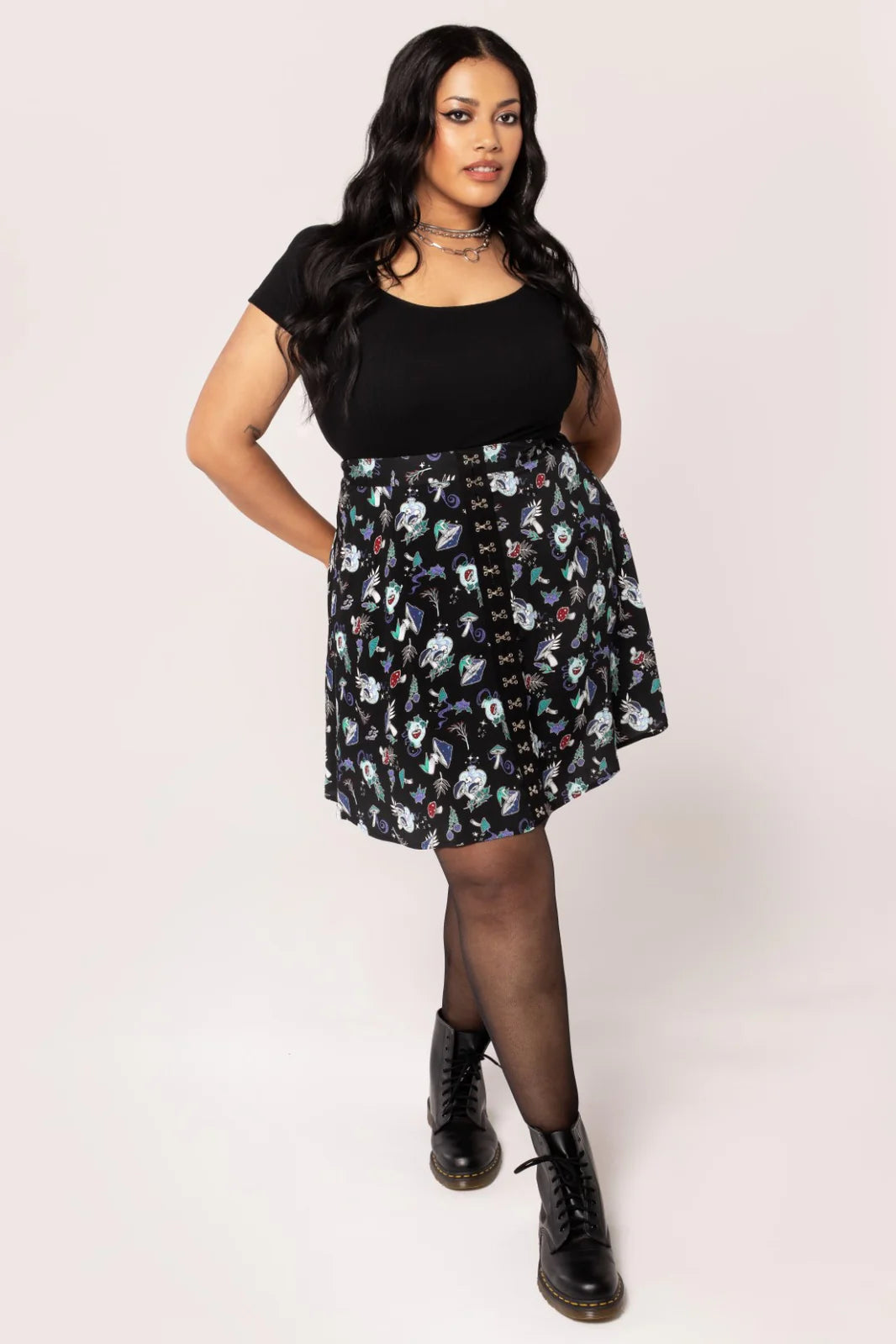 Woman with long dark hair wearing silver chain necklaces, a black top and the Amanita mushroom print mini skirt by Hell Bunny with black Doc Marten boots and sheer tights. She is standing with one foot in front of the other and her hands behind her back.