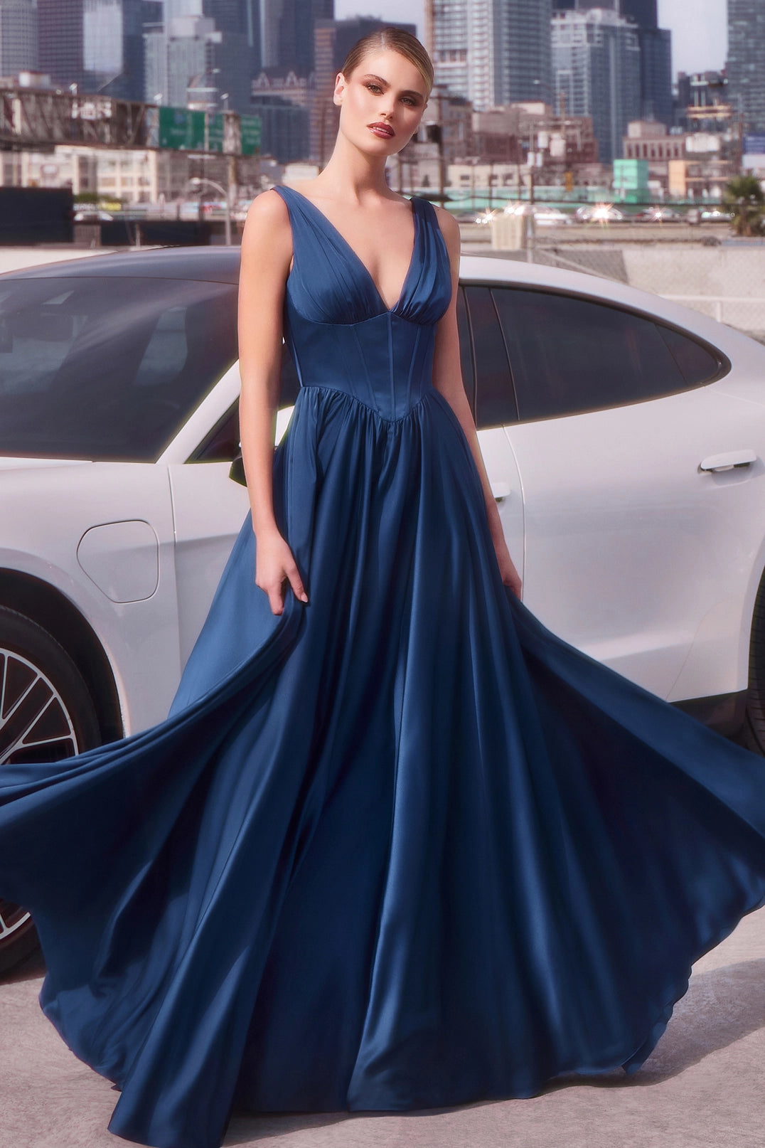 Woman in a blue evening gown standing next to a white car with a cityscape in the background