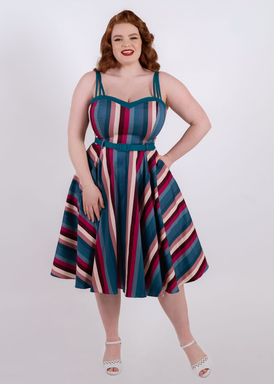 Redhead vintage pinup model smiling in red lipstick and a stripey blue, pink and white swing dress standing with one hand in her pocket