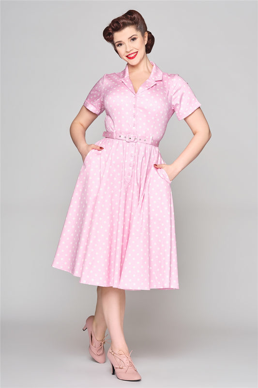 Happy, smiling brunette woman standing with her hands in her dress pockets. She is wearing a pink shirt dress with white polka dots all over and light pink heeled ankle boots.