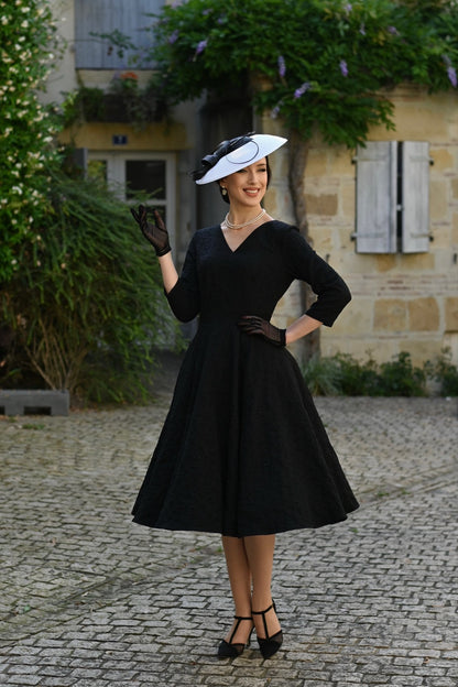 Woman in a black dress and hat standing on a cobblestone street with a building in the background.