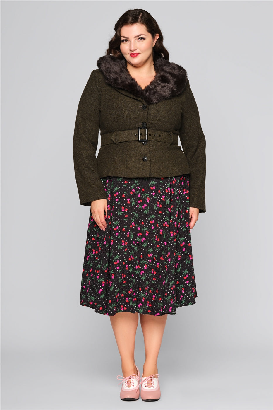 Curvy brunette model wearing a cherry print midi skirt, brogues and a classic olive green jacket with fur trim collar