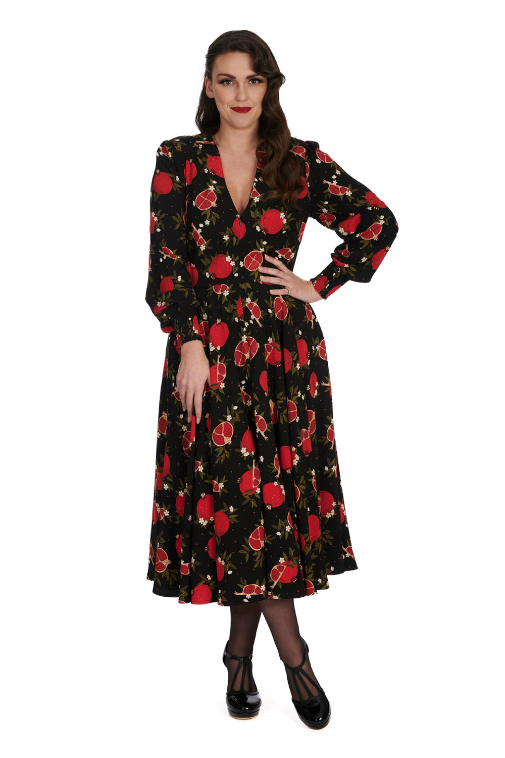 A woman wearing a long, black dress with a vibrant red pomegranate print. The dress features a V-neckline and full-length, slightly puffed sleeves gathered at the wrists. The skirt flows down to mid-calf, creating an elegant and vintage-inspired silhouette. The model pairs the dress with black stockings and shiny black T-strap heels, enhancing the retro vibe of the ensemble. She stands with one hand on her hip, confidently showcasing the dress's flattering fit.