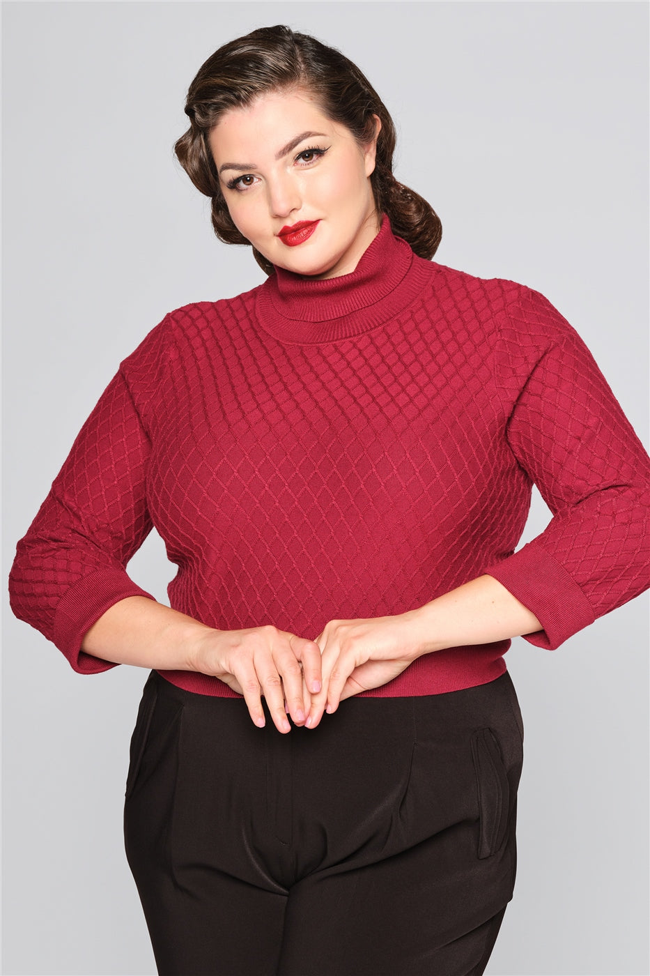Brunette curvy model with red lipstick holding her hands in front of her, wearing a burgundy turtleneck knitted jumper and high waisted black trousers