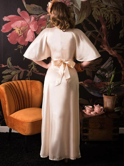 Glamorous woman wearing a floor length peach coloured satin lounging robe tie in a bow at the back, standing with her hands on her hips