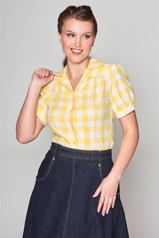 Smiling woman wearing a yellow and white blouse and denim skirt. She is holding one side of her collar. 