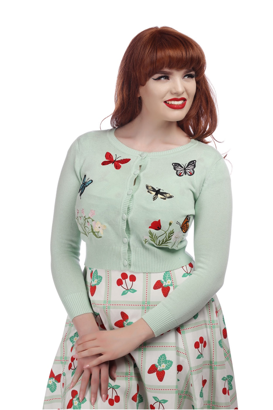 pretty vintage model smiling and wearing 50s inspired red lipstick and black eyeliner, a fruit print skirt and a mint green knitted cardigan with embroidered moths, butterflies and flowers